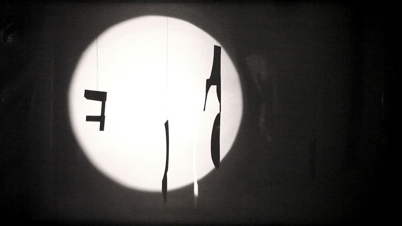Schwart-weißer Filmstill mit einem Leuchtkegel auf abstrake Formen