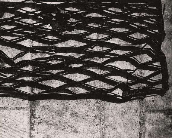 Die Fotografie Gitterstruktur von Helmut Hahn zeigt eine Nahaufnahme eines metallenen Gitters auf einem Steinboden